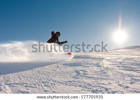 Teenage boy riding a snowboard downhill in sunny weather, spraying snow as he turns
