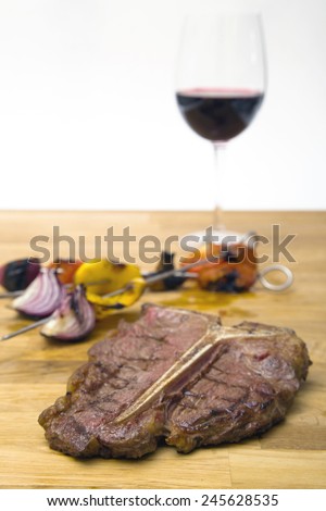 T bone steak on a wood plate with a glass of red wine in the background and some grilled vegetables on a spears
