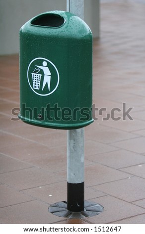 A green trash bin in a city. The Bin is fastened to a lightpole.