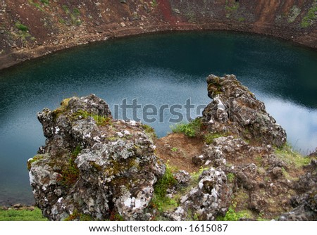 An old crater filled with water, lava in foreground jutting over a steep incline set against the deep blue water below.