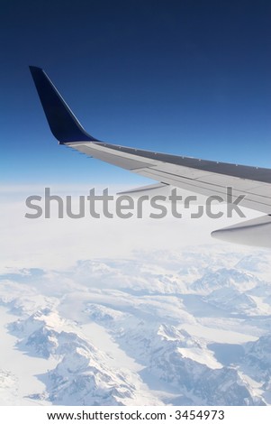 wing of a passenger jet at high altitude with snow covered mountain terrain in the mist below