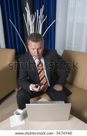 Businessman sitting in sofa texting on his mobile phone with coffee and laptop on table