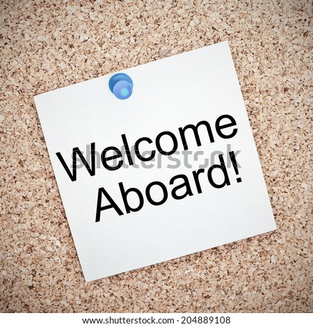 Welcome Aboard typed onto a yellow note and pinned to a cork notice board. A phrase used to welcome a new employee or team member.
