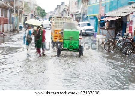 Defocussed view of flash flood at Indian city street with auto rickshaw, bicycles and pedestrians after monsoon rain. India, Tamil Nadu