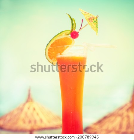 Tequila sunrise cocktail with fruits  decoration at tropical ocean beach with umbrella. Vintage style, hipster colors image with copy space for party invitation text