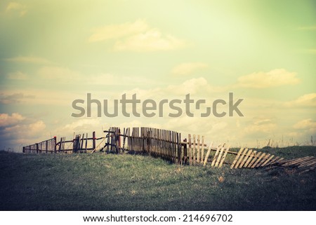 Abandoned countryside. Concept summer landscape with old broken fence at pasture under cloudy sky. Nature background in vintage style