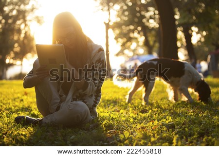Girl Using Tablet In The Park On The Grass With The Dog At Sunset