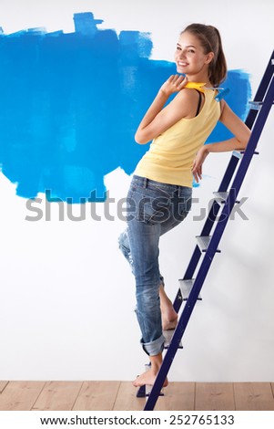 Beautiful young woman doing wall painting, standing on ladder