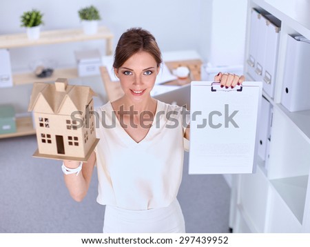 Portrait of female architect holding a little house, standing in office .