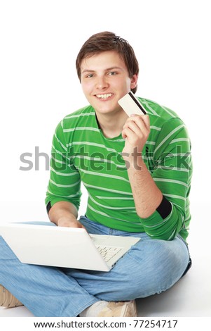 A young man sitting on the floor with a laptop, holding a credit card