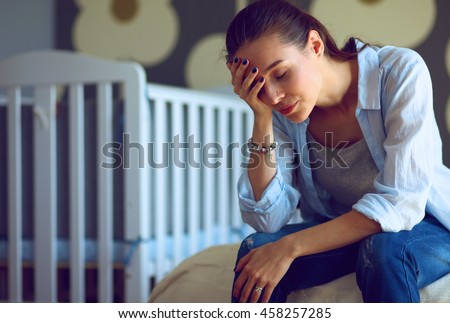 Young tired woman sitting on the bed near children\'s cot.