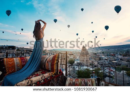 Young beautiful woman wearing elegant long dress in front of Cappadocia landscape at sunshine with balloons in the air. Turkey.