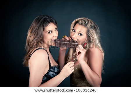 Two smiling attractive girlfriends eating chocolate bar, blond and brunette on dark background.