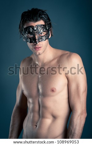 Portrait of young man shirtless with carnival mask against dark background.