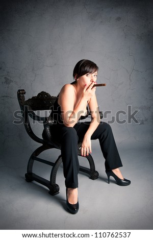 Woman smoking cigar sit on an old black chair. Studio fashion photo with grunge dark background.