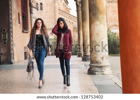 Two girls walking outdoors in Saint Stephen square, Bologna, Italy. Full body.