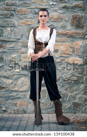 Beautiful girl wearing a medieval dress with sword. Stone wall background.