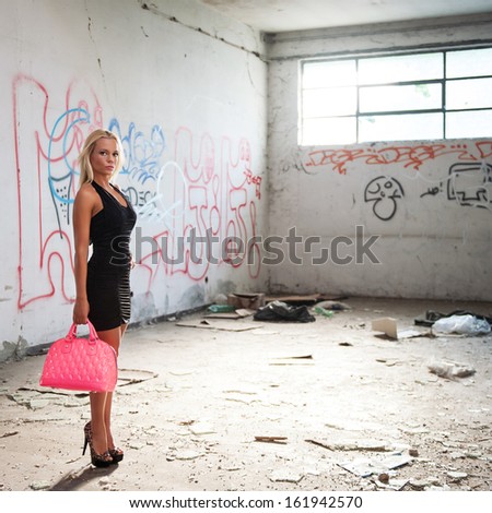 Sensual blonde woman portrait with black dress and pink bag inside abandoned factory. Full body.