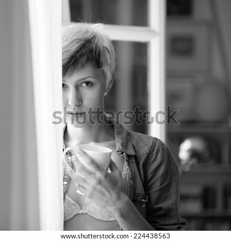 Intimate portrait of beautiful woman drinking a cup of tea in front of window at home. Black and white image.