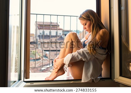 Sensual young blonde woman portrait wearing lingerie at the window in hotel room. Filtered image.