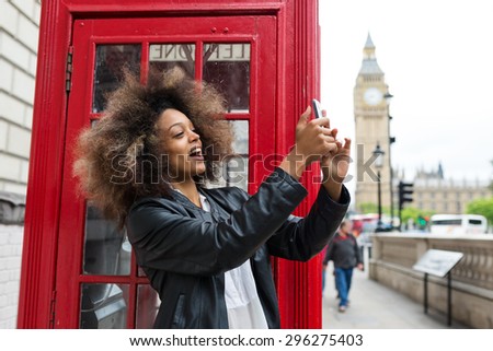 Young woman portrait close to red telephone box in London taking a selfie with smart phone. Big Ben in the background.