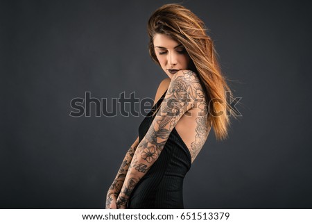 Intimate woman studio portrait with long black dress and tattoos against dark background.