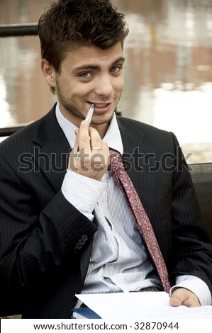 Young man in a suit sits outside and bites a pen