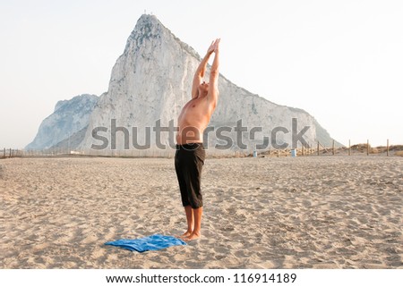 Mature man practising yoga at the beach with the rock of gibraltar as background exercising for balance of body and mind