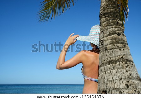 A woman standing under a palm tree watching the ocean.
