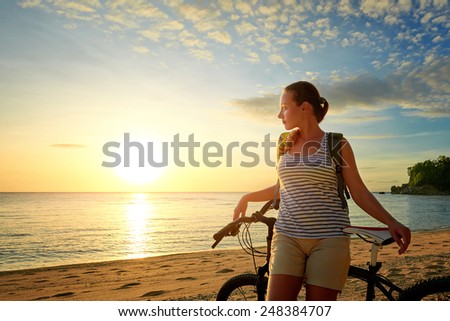 Young woman with backpack standing on the shore near his bike and looking on sunset. Traveling along Asia, active lifestyle concept