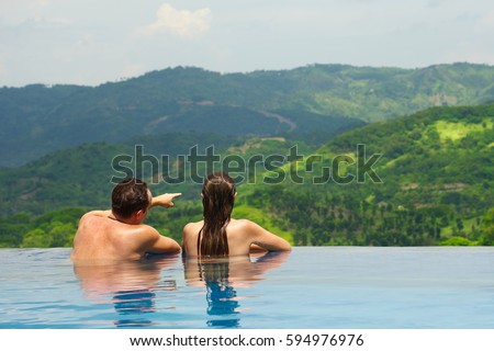 Rear view of happy couple in the pool looking at mountain landscape.\
Enjoying beautiful mountains. Man, woman together on summer travel to luxury resort. Summertime relax.