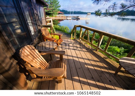 Muskoka chairs sitting on a wooden porch facing a calm lake. In the background there are Adirondack and Muskoka chairs on a wooden dock