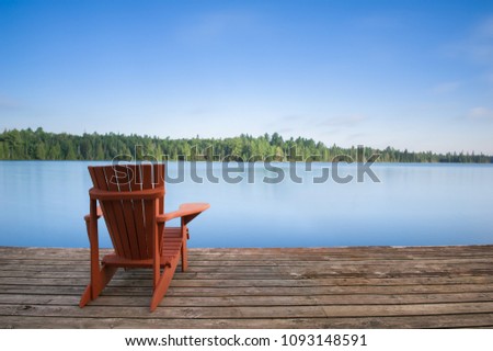 Adirondack chair sitting on a wood dock facing a calm lake. Across the water there are green trees.