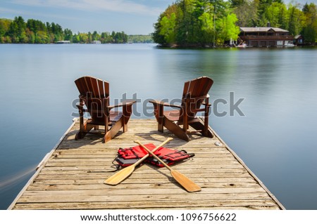 Two Muskoka chairs sitting on a wood dock facing a lake. Across the calm water is a brown cottage nestled among green trees. Some life jackets and canoe paddles are on the dock.