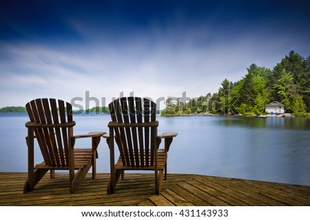 Two Muskoka chairs sitting on a wood dock facing a lake. Across the calm water is a white cottage nestled among green trees. There is a boat dock on the water in front of the cottage.