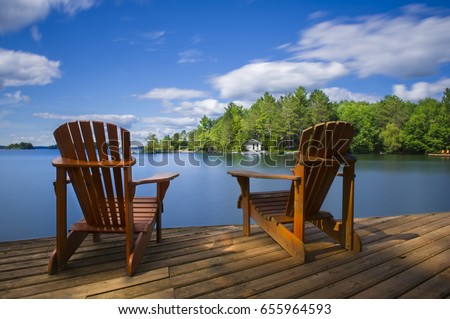 Two Muskoka chairs sitting on a wood dock facing a calm lake. Across the water is a white cottage nestled among green trees. There is a boat dock on the water in front of the cottage.