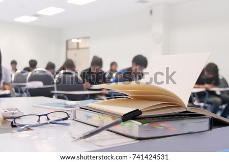 A notebook/pen/books/grasses of teacher on table and students during study or quiz, test and exam in large lecture room / University classroom. Students are in uniform classroom educational school.