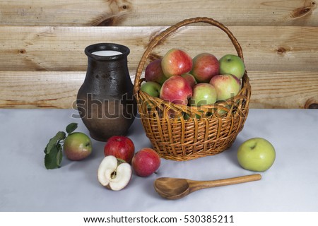 Still life with ripe apples in an openwork wicker basket and clay jug with milk on the background of the log wall.