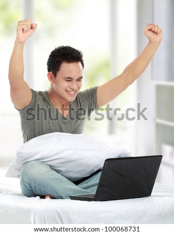 A young man sitting on a bed with laptop celebrating success