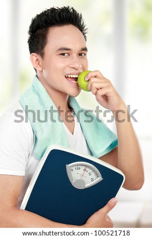 healthy fitness man carrying a weight scale while eating fresh green apple