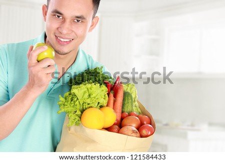 smiling young man holding shopping bag full of groceries isolated against white background