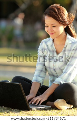 Young female sitting cross-legged on grass with laptop and books doing a homework