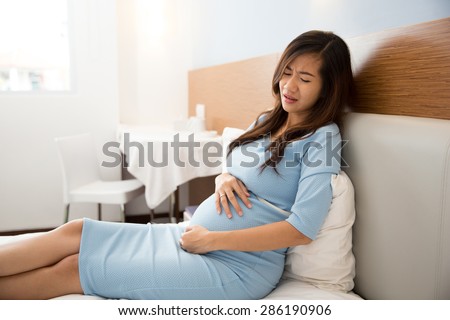 A portrait of an Asian pregnant woman has stomachache sitting on her bed