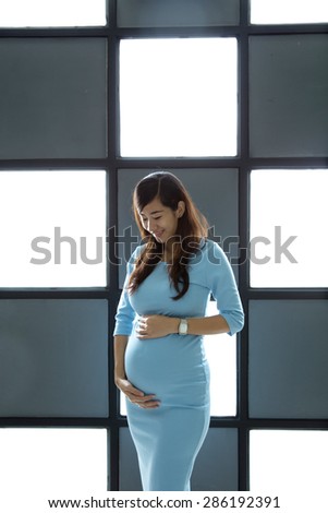 A portrait of an Asian pregnant woman standing infront of a light board, smiling look at her tummy