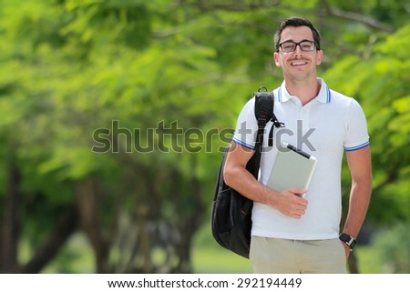 portrait of smiling handsome college student with backpack holding a tablet and hand in pockets with copy space