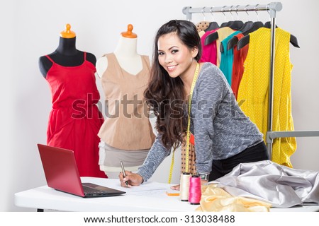 A portrait of a young asian designer woman using a laptop and smiling,clothes hanged as background