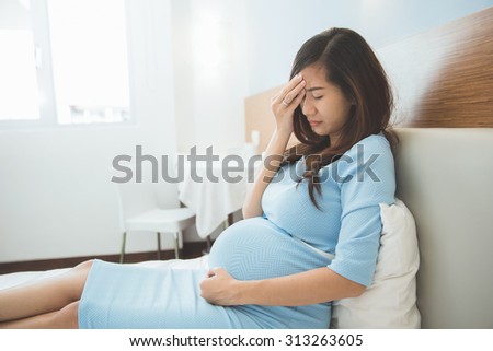 A portrait of an Asian pregnant woman has headache sitting on her bed