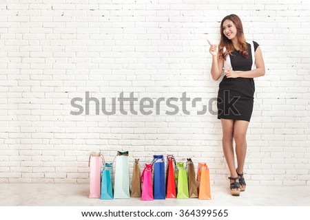 full body portrait of beautiful young woman standing next to shopping bags while pointing at copy space on white brick wall background