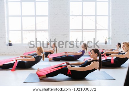 Group of six women are doing stretching exercise with red resistance bands in white studio interior. Teamwork, good mood and healthy lifestyle concept.