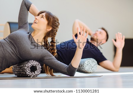 Gorgeous woman instructor performing back exercise on a foam roller at same time with handsome guy at pilates studio. Coach and patient doing fascia exercise on side surface of back.